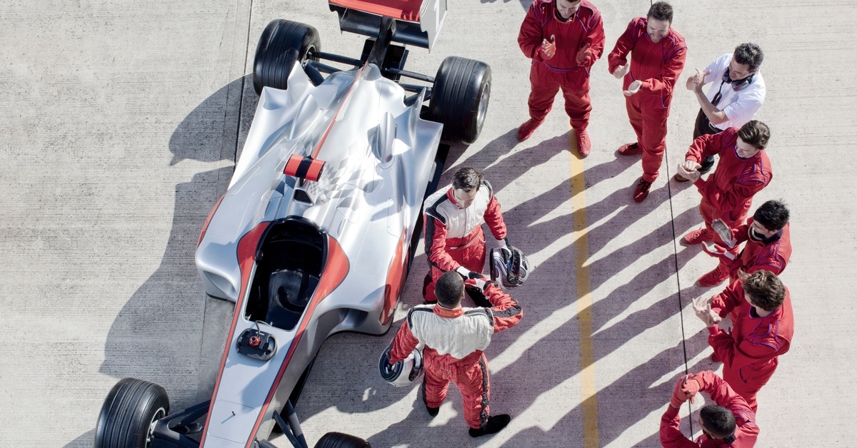 A racing team wearing red racing suits surrounding a silver and red formula-style race car parked on a concrete pit lane.