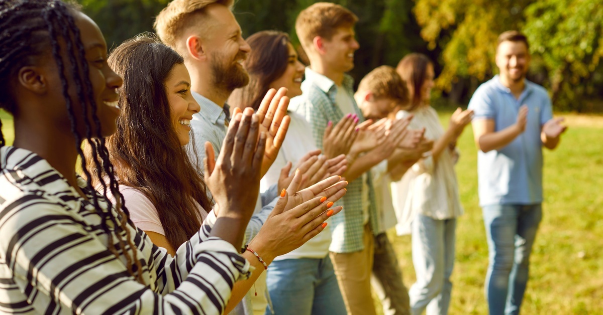 A long line of people standing in an open field, all smiling and clapping together. There are a lot of trees in the background.