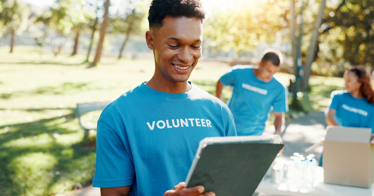 A man wearing a blue volunteer shirt, looking at a clipboard while smiling. There are people setting out water behind him.