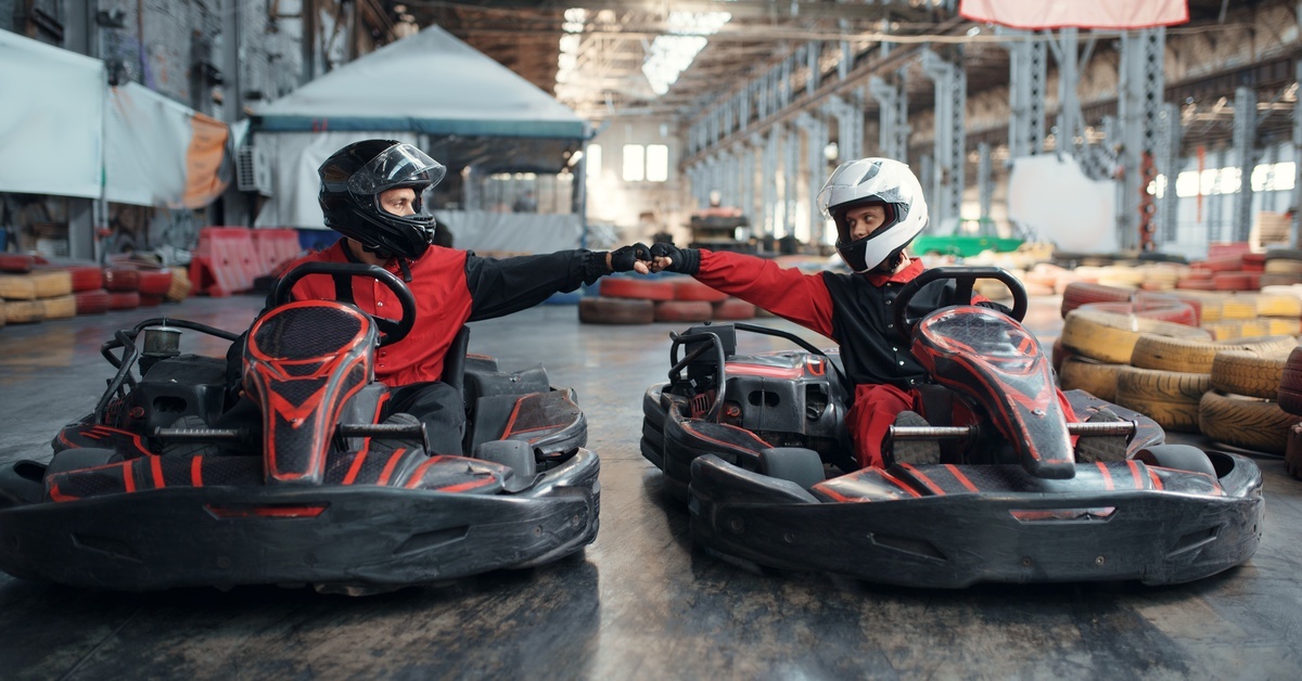 Two guys sitting in go-karts, reaching out and fist bumping each other. Their suits and karts are mostly black and red.