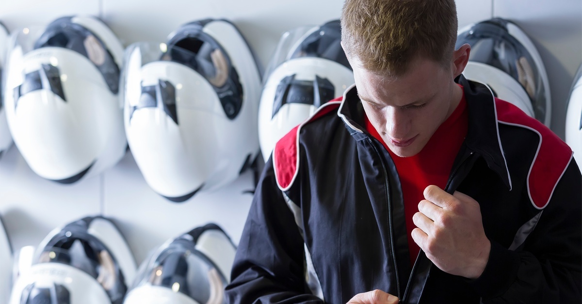 A man looking down while zipping up his racing suit. He is standing in front of a wall full of white racing helmets.